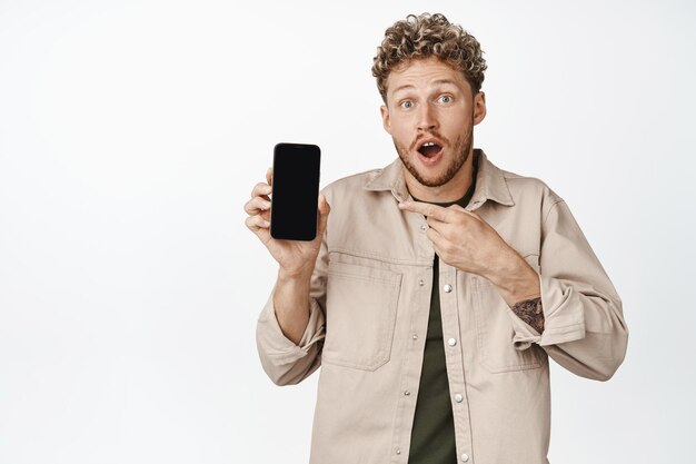 Возбужденный блондин кудрявый парень, указывающий пальцем на экран мобильного телефона, выглядит удивленным, показывая потрясающие новости онлайн, стоя на белом фоне