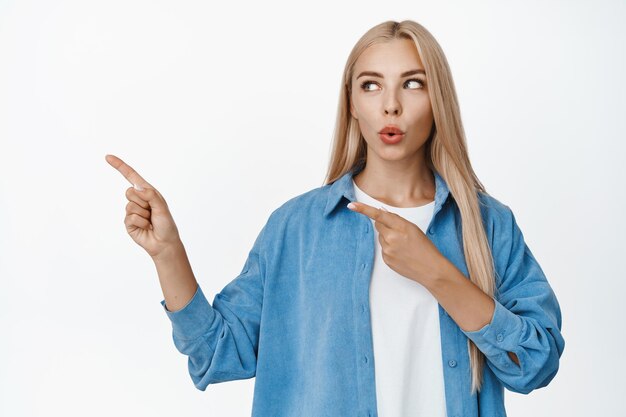 Excited blond girl in casual clothes pointing and looking left at announcement showing advertisement standing against white background