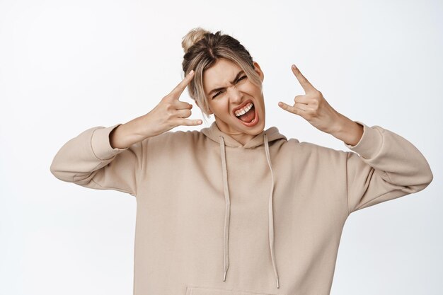 Excited blond girl enjoying event shows heavy metal horns rock gesture and yelling sassy standing against white background