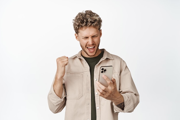 Excited blond man looking at mobile phone and triumphing watching video and winning bet makng fist pump achieve goal on smartphone standing over white background