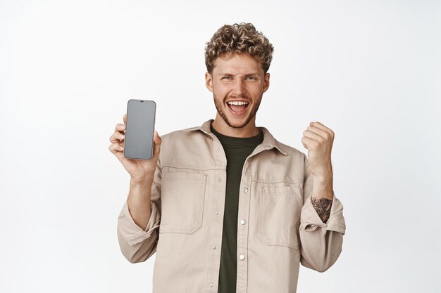 興奮した金髪の男が携帯電話の画面を表示し、アプリの勝利と白い背景に立っているスマートフォンのデモンストレーションで楽しい勝利のお金を叫ぶ