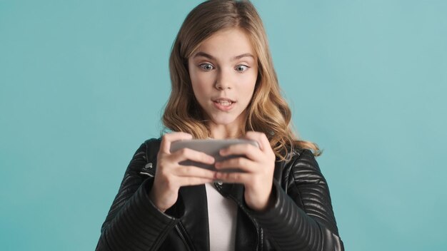 Excited blond teenager girl playing on smartphone being obsessed with online games looking nervous over blue background Gamer girl