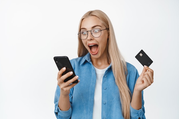 Foto gratuita eccitata donna bionda con gli occhiali che guarda lo schermo dello smartphone e urla, fissando sorpresa e stupita sul bianco