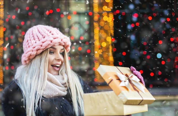 興奮した金髪の女性は、冬のフェアで贈り物を楽しんでいるピンクのニット帽子をかぶっています。テキスト用のスペース Premium写真