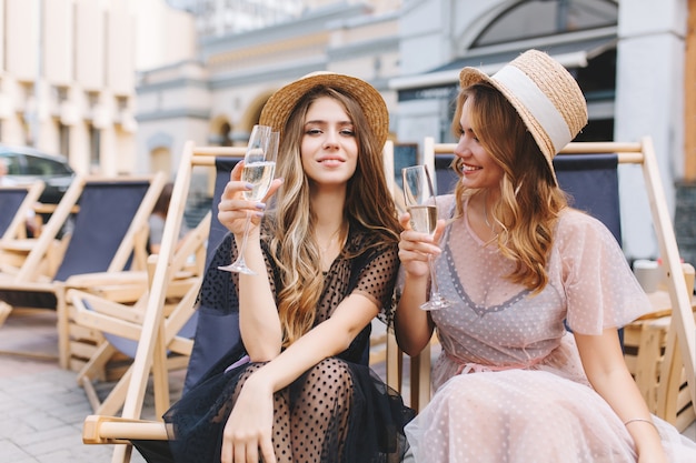 Excited blonde girl in transparent white dress looking at sister which posing holding wineglass