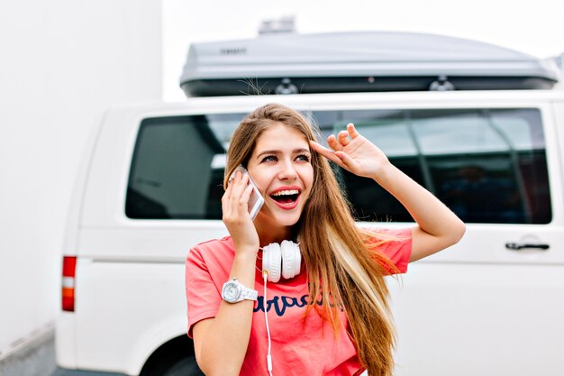 Excited blonde girl wearing pink shirt and white headphones talking on phone with friend and looking in distance