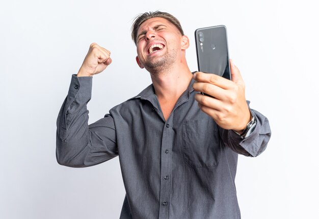 Excited blonde handsome man holding phone and keeping fist up