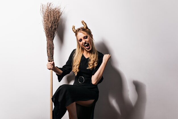 Excited blonde witch posing with broom. Beautiful vampire in black dress expressing happiness in halloween.