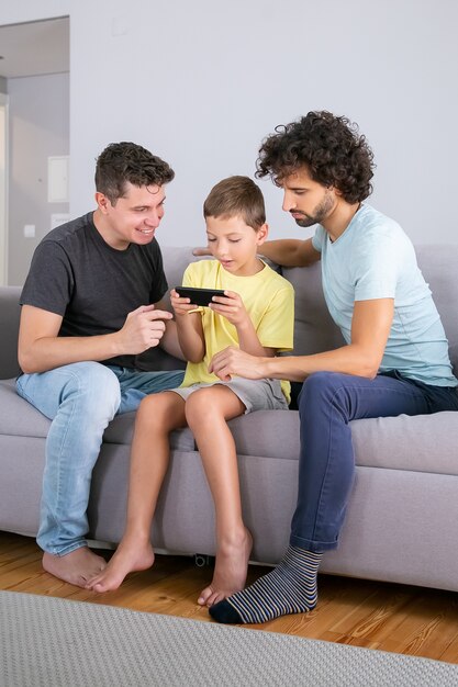 Взволнованный мальчик играет в игру на мобильном телефоне. Два папы помогают сыну использовать онлайн-приложение на мобильном телефоне. Вертикальный снимок. Семья дома и концепция коммуникации