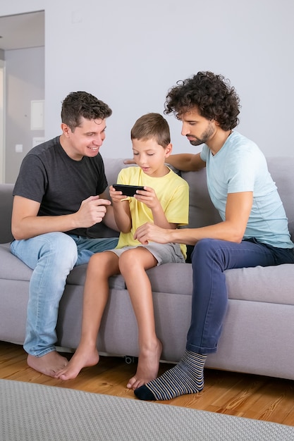 携帯電話でゲームをしている興奮した少年。息子がセルでオンラインアプリを使用するのを手伝っている2人のお父さん。垂直ショット。家庭での家族とコミュニケーションの概念