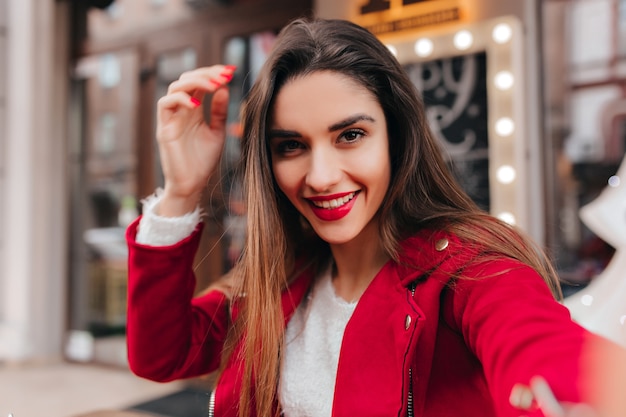 Excited brown-haired girl with stylish makeup making selfie on blur urban space