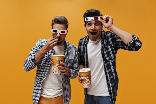 Excited brunet man puts off 3D eyeglasses Friends in blue checkered shirts eat popcorn and watch movie on orange background