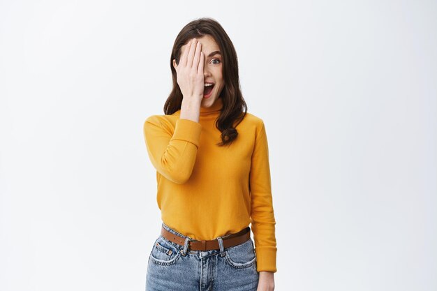 Excited brunette girl cover half of face with palm and scream with excitement and happiness, checking out something cool, standing happy on white background