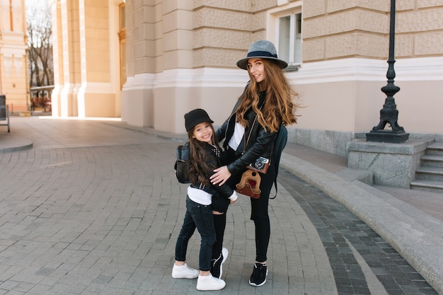 Excited brunette girl in hat and stylish jacket embracing her mother's leg standing in the middle of street.