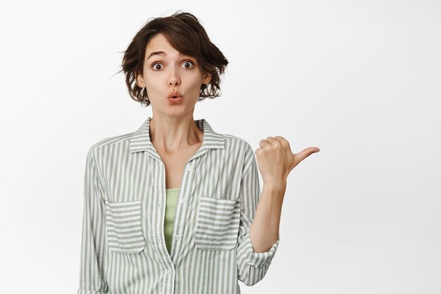 Excited brunette girl looks interested, points right, asks question about banner or product you place on empty copy space, stands against white background.