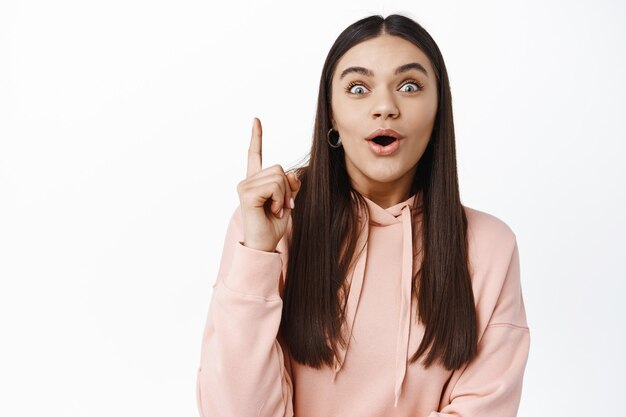 Excited brunette girl pitching an idea, raising finger up and gasping amazed, saying interesting suggestion, found solution or made-up plan, standing against white wall