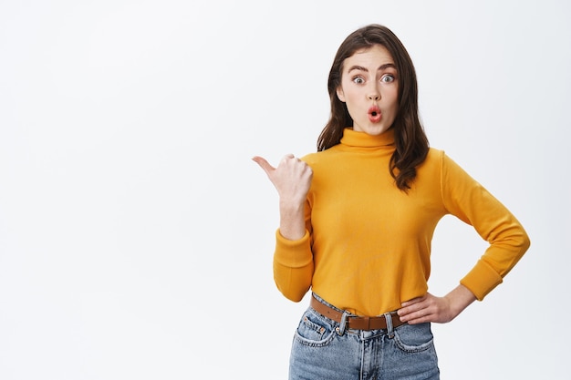 Excited brunette girl pointing finger left at promo deal, showing banner with amused face, standing against white wall