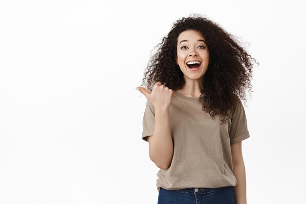 Excited brunette girl showing advertisement pointing thumb aside and smiling amazed recommending product or store click on link standing over white background