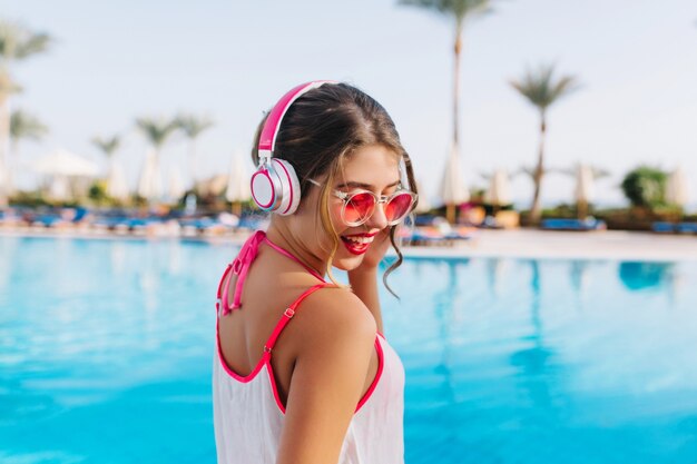 Excited brunette girl with tanned skin listening favoring music while sunbathing by the open-air pool.