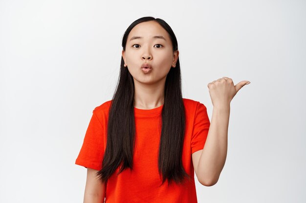 Возбужденная брюнетка кореянка показывает объявление, указывая пальцем вправо, и выглядит удивленной, говоря, вау, стоя на белом фоне