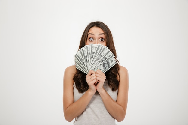 Free photo excited brunette woman covering her face with fan of dollar banknotes, expressing shock and surprise over white wall