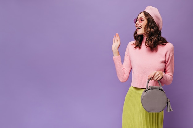 Excited brunette woman in green skirt, pink beret and sweater waves hand in greeting