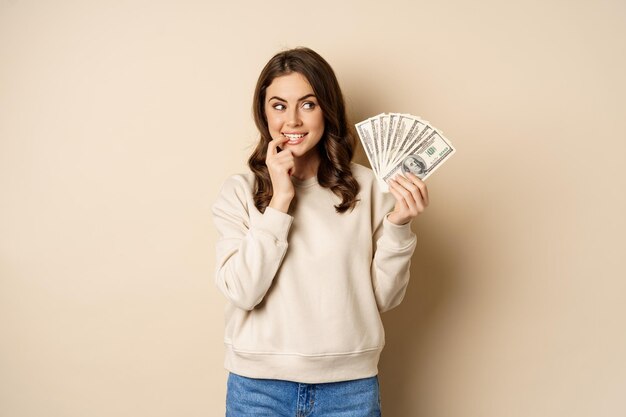 Excited brunette woman holding money and biting finger with yearning face watns to buy smth shopohol...