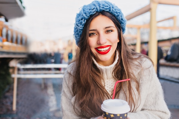 흐림시에 차를 마시는 갈색 눈을 가진 흥분된 갈색 머리 여자. 추운 날에 뜨거운 커피 한잔 들고 코트와 파란색 모자에 화려한 검은 머리 아가씨의 야외 사진.