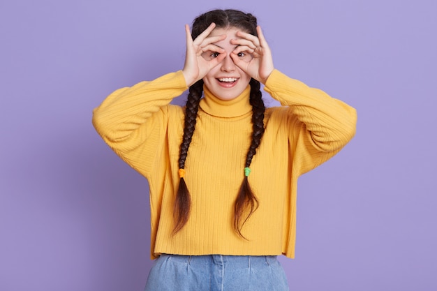excited brunette young woman with two pigtails standing with happy expression and covering her eyes with ok signs