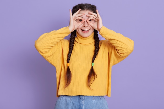 Excited brunette young woman with two pigtails standing with happy expression and covering her eyes with ok signs