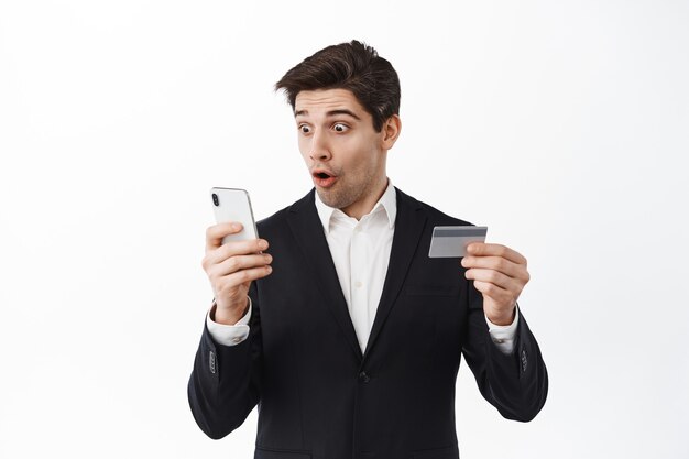 Excited business man in suit look at his phone, reading screen with amazed face and holding credit card, log in to online banking app, standing over white wall