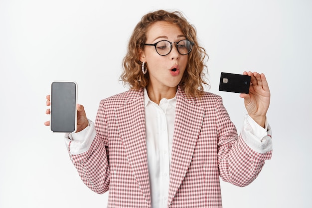 Excited business woman in glasses, shows mobile phone screen, looks at credit card with amazed face expression on white