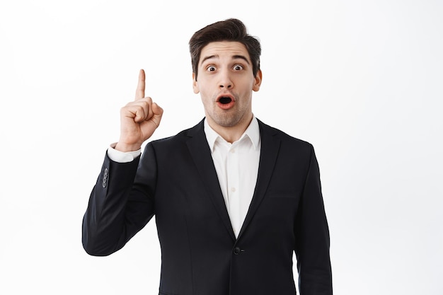 Excited businessman raise index finger, pitching an idea, think up solution, perfect plan, say suggestion, standing over white background in black suit