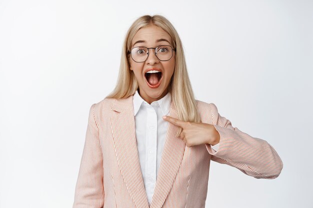 Excited businesswoman in glasses and suit pointing finger at herself employee got a job looking surprised at camera reaction to promotion white background