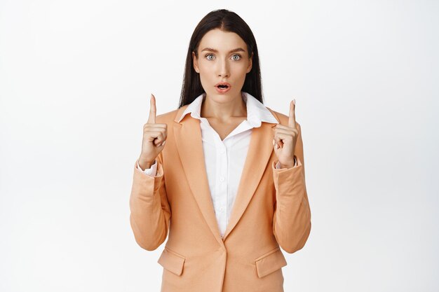 Excited businesswoman pointing fingers up looking impressed at camera standing in suit over white background