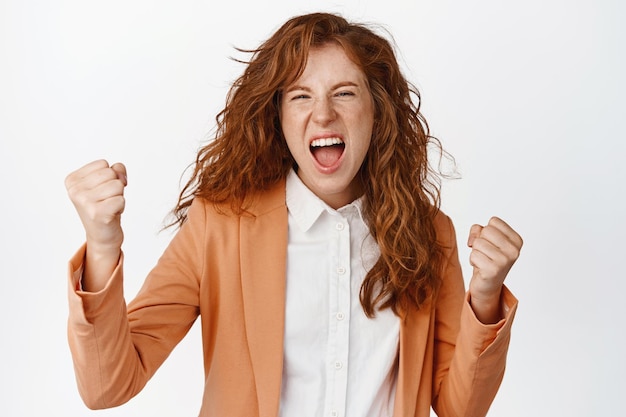 Excited businesswoman screaming Saleswoman triumphing with clenched fists cheering and looking happy standing in suit over white background