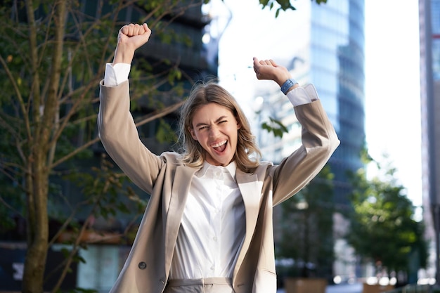 Free photo excited businesswoman screams and celebrates lifts hands up does hooray gesture celebrates victory achieve goal stands on street