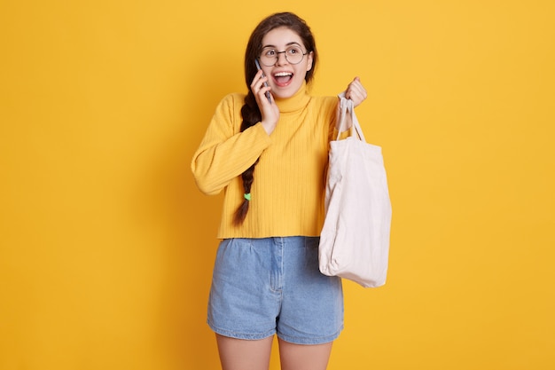 Excited buyer girl with bag in hands talking to phone, keeping mouth widely opened, wearing stylish sweater, short and glasses