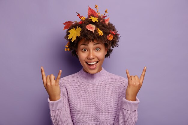 Excited carefree Afro woman has fun during autumn party, makes rock n roll gesture, heavy metal sign, has curly hairstyle with autumnal foliage, feels energized, wears purple casual sweater.