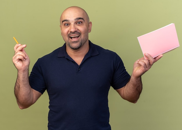 Excited casual middle-aged man looking at front raising pencil and note pad isolated on olive green wall