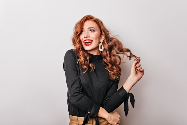 excited caucasian girl in black blouse playing with her ginger hair.appealing young woman wears golden earrings.