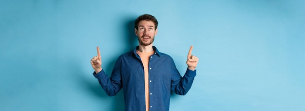 Foto gratuita eccitato ragazzo caucasico che guarda e punta le dita verso l'alto mostrando pubblicità in piedi sul backgro blu