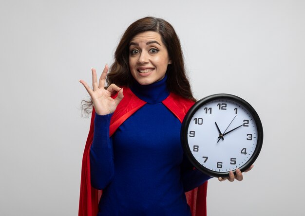 Excited caucasian superhero girl with red cape gestures ok hand sign and holds clock isolated on white wall with copy space