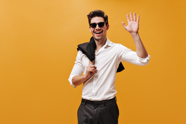 Возбужденный очаровательный брюнет в белой рубашке, солнцезащитных очках и черных брюках улыбается, держит куртку и машет рукой в приветствии на оранжевой стене.