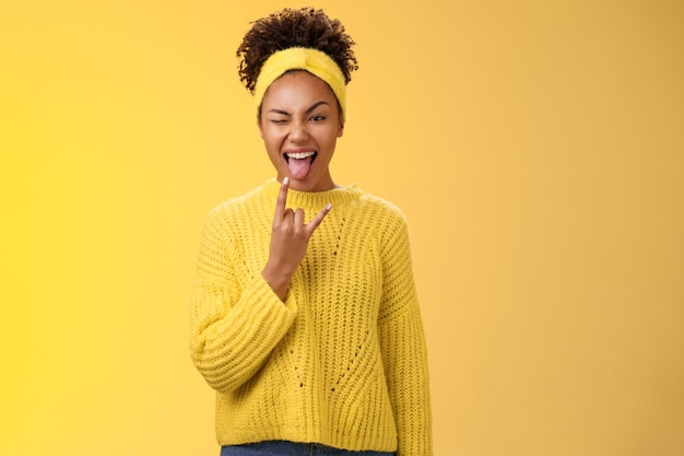 Excited charming smiling daring sassy young african american woman having fun show tongue rock-n-roll heavy metal gesture winking happily enjoying cool awesome party, standing yellow background.