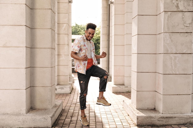 Excited charming young man in black stylish jeans and floral shirt rejoices smiles and dances outdoors