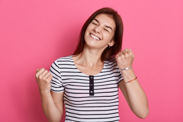 Excited cheerful female with joyful expression, cheers and clenches fists, celebrating her success, poses against pink wall, dresses casual white and black striped t shirt.