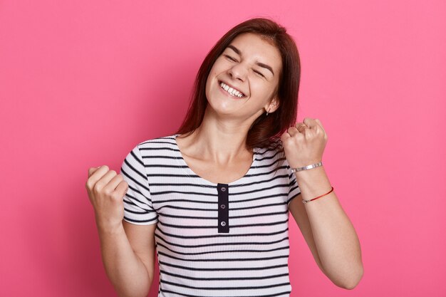 Excited cheerful female with joyful expression, cheers and clenches fists, celebrating her success, poses against pink wall, dresses casual white and black striped t shirt.