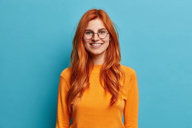 Excited cheerful redhead woman with sincere toothy smile has fun and looks directly wears round optical glasses dressed in orange jumper.