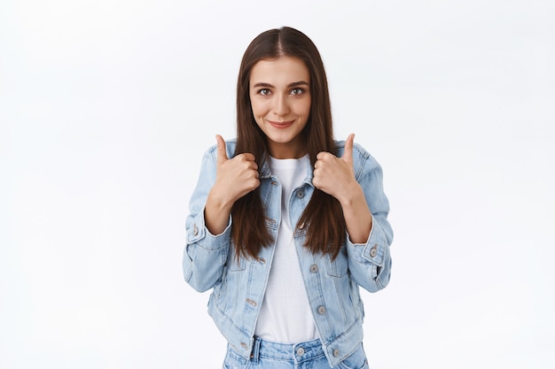 Excited, cheerful stylish caucasian female brunette in denim jacket, showing thumbs-up and smiling delighted, approve amazing idea and nod in agreement, like nice offer, white background